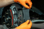 GrimmSpeed titanium front shim being fitted behind a pad in a 4-piston caliper (product illustration) - GrimmSpeed Thumbnail
