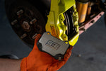 GrimmSpeed titanium rear shim being fitted behind a pad in a Subaru WRX STI yellow Brembo 2-piston rear caliper - GrimmSpeed Thumbnail